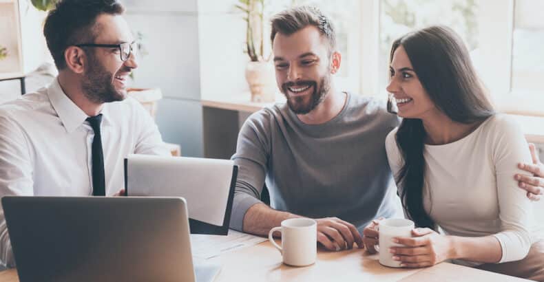 A couple sit with a financial advisor. He is holding papers and there is a laptop open on the desk. Everyone has a happy expression.
